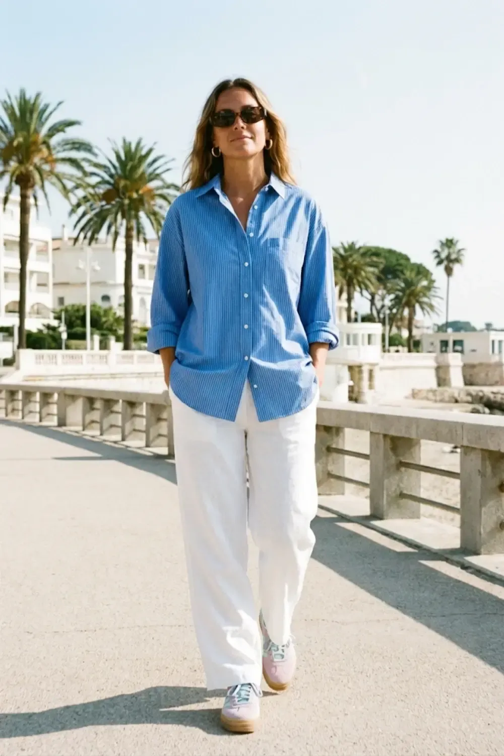 A woman walking along a sunny promenade with palm trees in the background. She completes the coastal-chic look with Adidas Gazelle Bold sneakers and dark sunglasses.