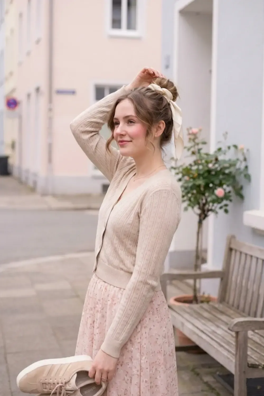 A smiling woman standing on a quiet street. She is fixing a messy bun which is tied with a large, cream-colored satin ribbon and holding Adidas Gazelle Bold sneakers