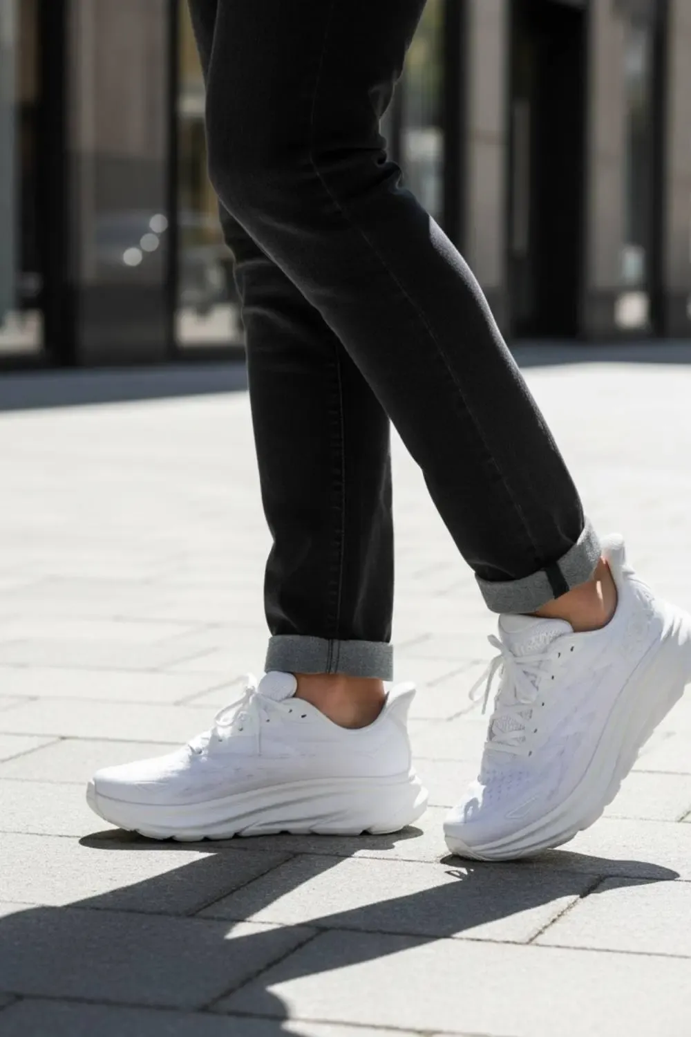 A man wearing white Hoka Clifton sneakers paired with dark denim jeans for a casual men's fashion look.