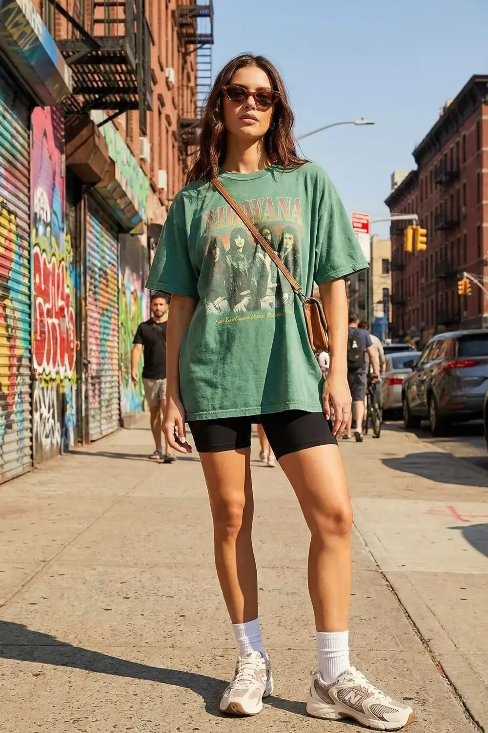 Woman in a green Nirvana T-shirt and black biker shorts wearing New Balance 530 sneakers on a graffiti-filled city street.