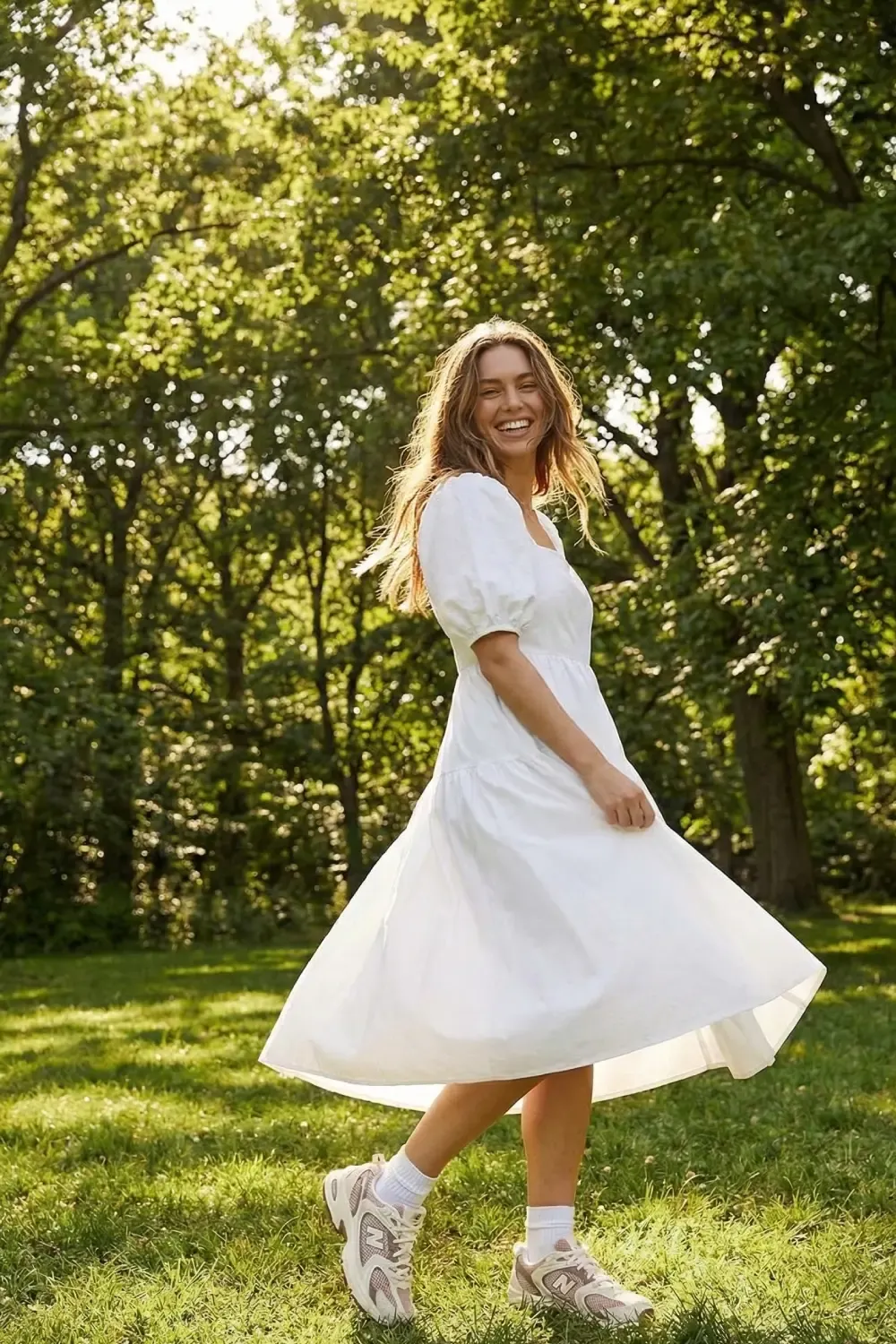 Woman in a white summer dress wearing New Balance 530 sneakers while smiling and walking in a sunny park.