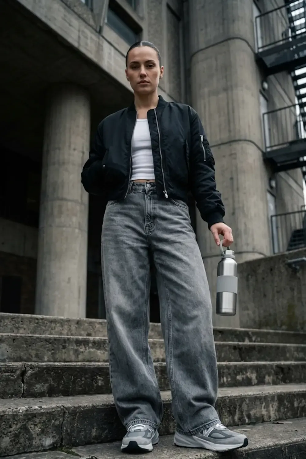 A woman standing outside a brutalist concrete building, wearing oversized wide-leg 'dad' jeans and a structured look with New Balance 990v6