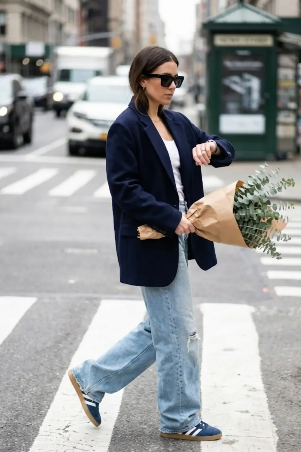 A woman crossing a city street, wearing extra-long light-wash wide-leg jeans that 'puddle' slightly at the bottom with Adidas Gazelle
